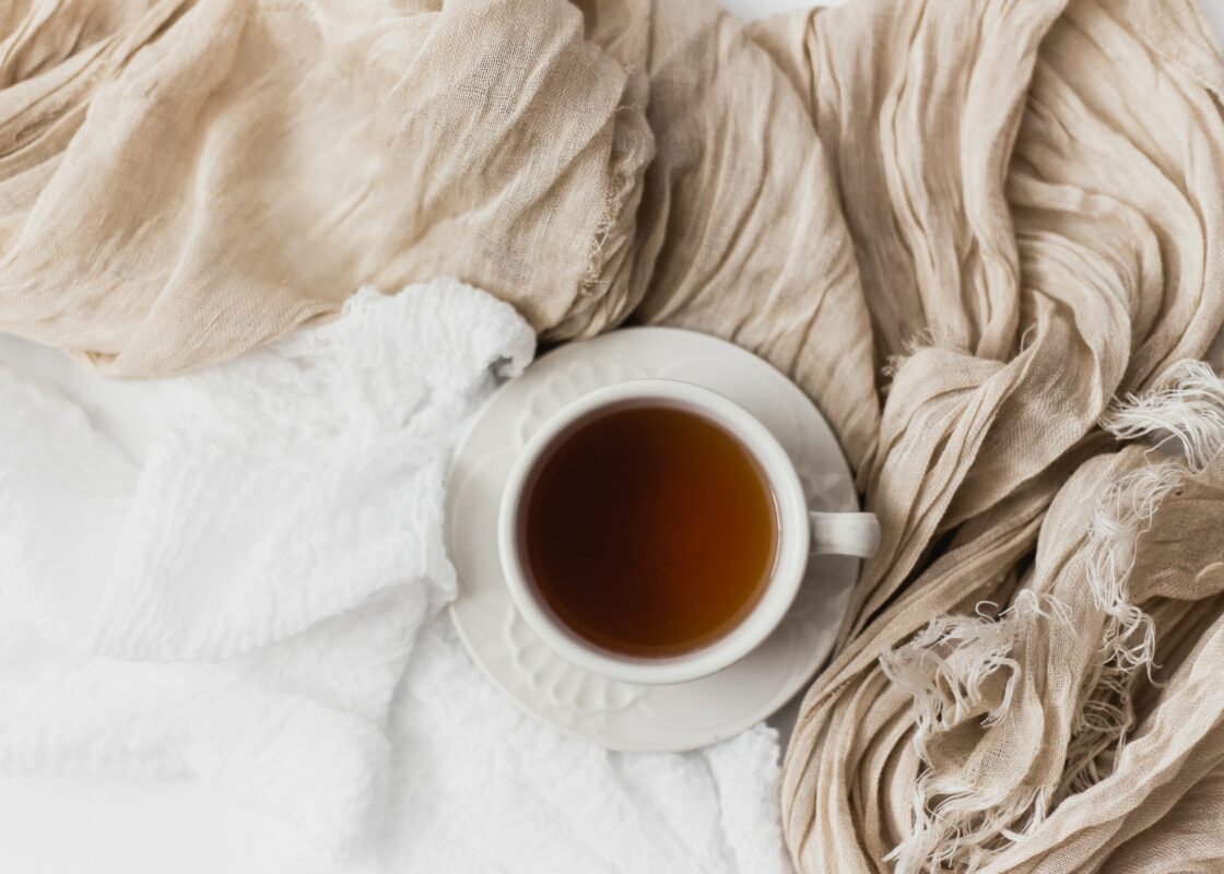 Eine Tasse Tee mit dunkler Flüssigkeit auf einer Untertasse steht inmitten weicher, strukturierter beiger und weißer Stoffe und schafft eine gemütliche und warme Atmosphäre, während die Winterkälte naht.