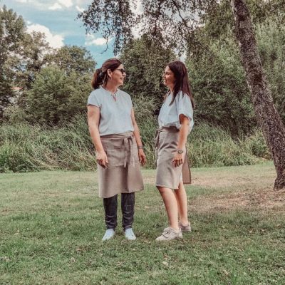 Zwei Menschen mit Schürzen stehen auf einer Rasenfläche mit Bäumen im Hintergrund. Sie stehen sich lächelnd gegenüber, unter einem leicht bewölkten Himmel.