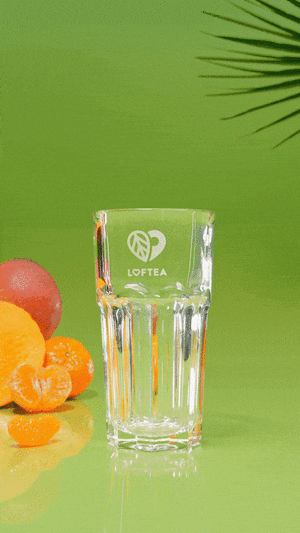 Ein Glasbecher mit dem Logo LOFTEA füllt sich mit Eistee vor einem grünen Hintergrund. Frische Orangenscheiben und ein Palmblatt im Hintergrund sorgen für eine erfrischende, tropische Szene.