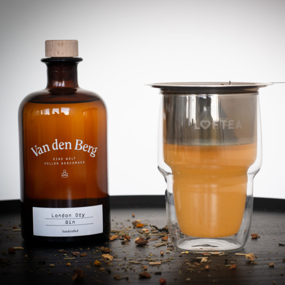 Ein VDB x LOFTEA GIN SET mit Doppelwandglas besteht aus einer braunen Flasche mit der Aufschrift Van den Berg London Dry Gin neben einem doppelwandigen Glas mit bernsteinfarbener Flüssigkeit. Auf dem Glas sitzt ein Metall-Sieb, umgeben von verstreuten LOFTEA-Blättern auf einer glatten schwarzen Oberfläche.