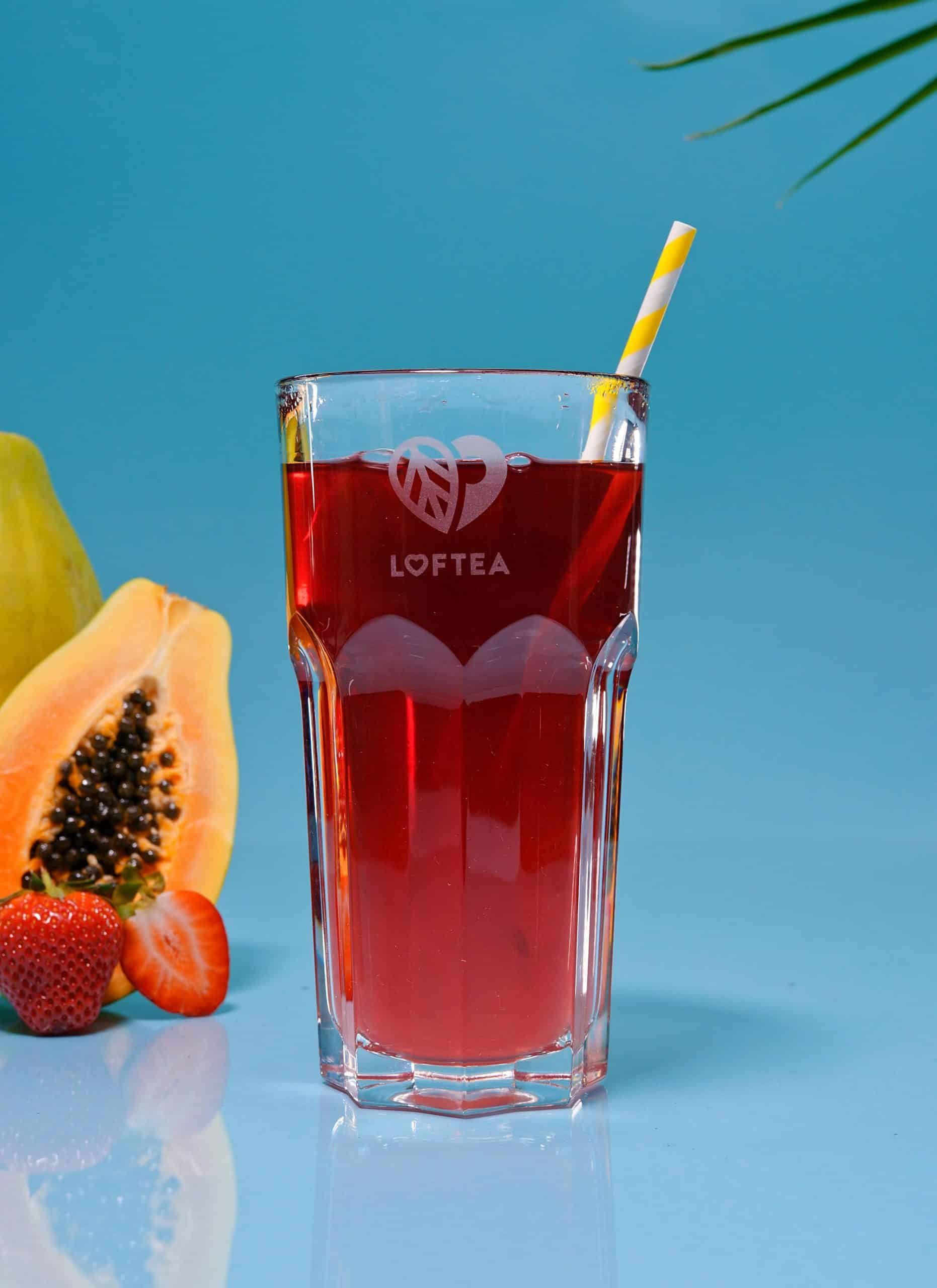 Ein hohes Glas mit erfrischendem ELLI ERDBEER, mit einem gelb-weiß gestreiften Strohhalm der Marke Loftea. Um das Glas herum befinden sich eine geschnittene Papaya, Erdbeeren und eine Guave auf einem leuchtend blauen Hintergrund.