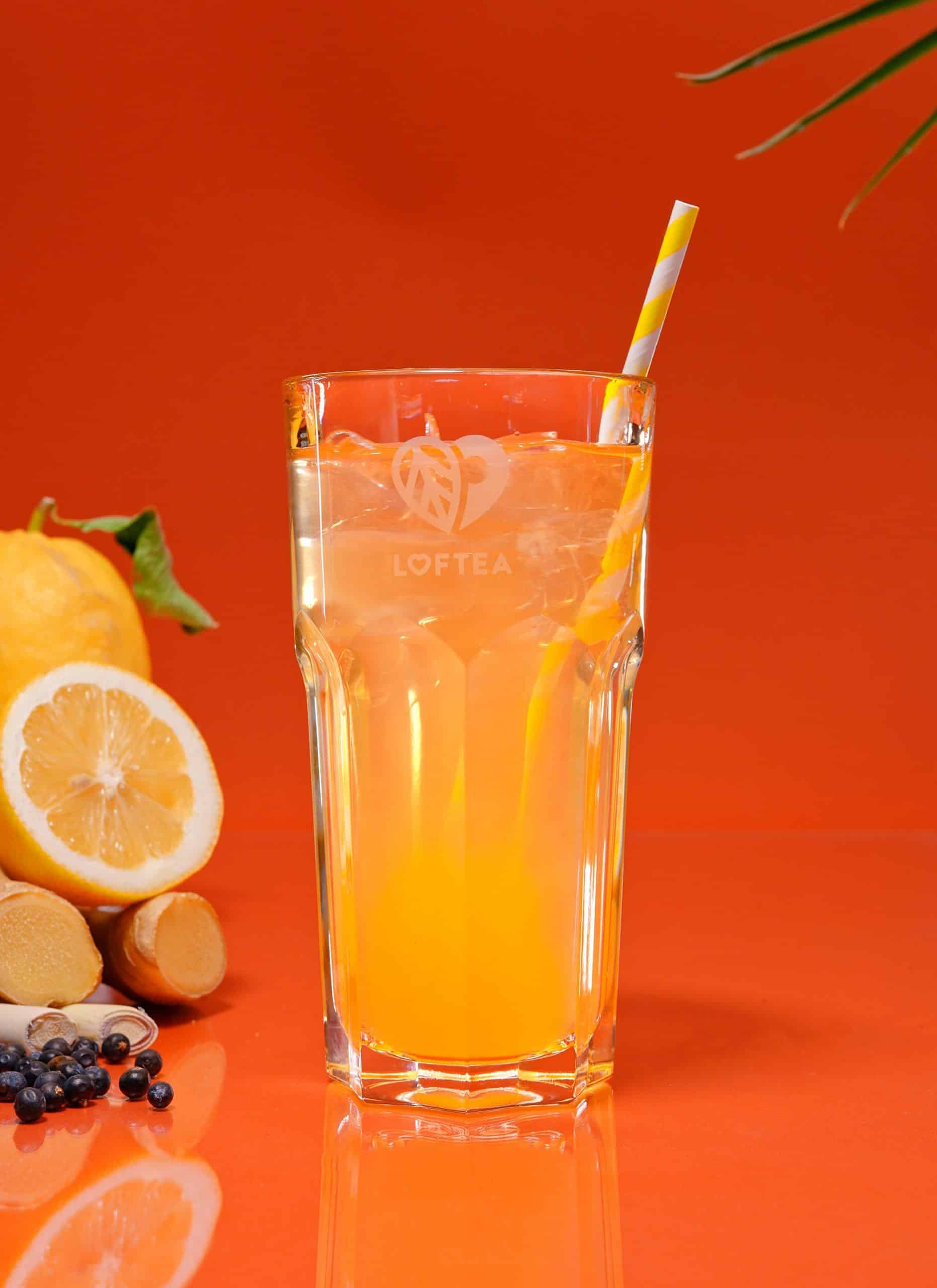Ein großes Glas Orangen-Eistee mit gestreiftem Strohhalm steht vor einem orangefarbenen Hintergrund. Mit einem Spritzer GIN LOFER angereichert, sind frische Zitronenscheiben, Ingwer und Beeren kunstvoll um das Glas herum angeordnet und verstärken die lebendige Komposition.
