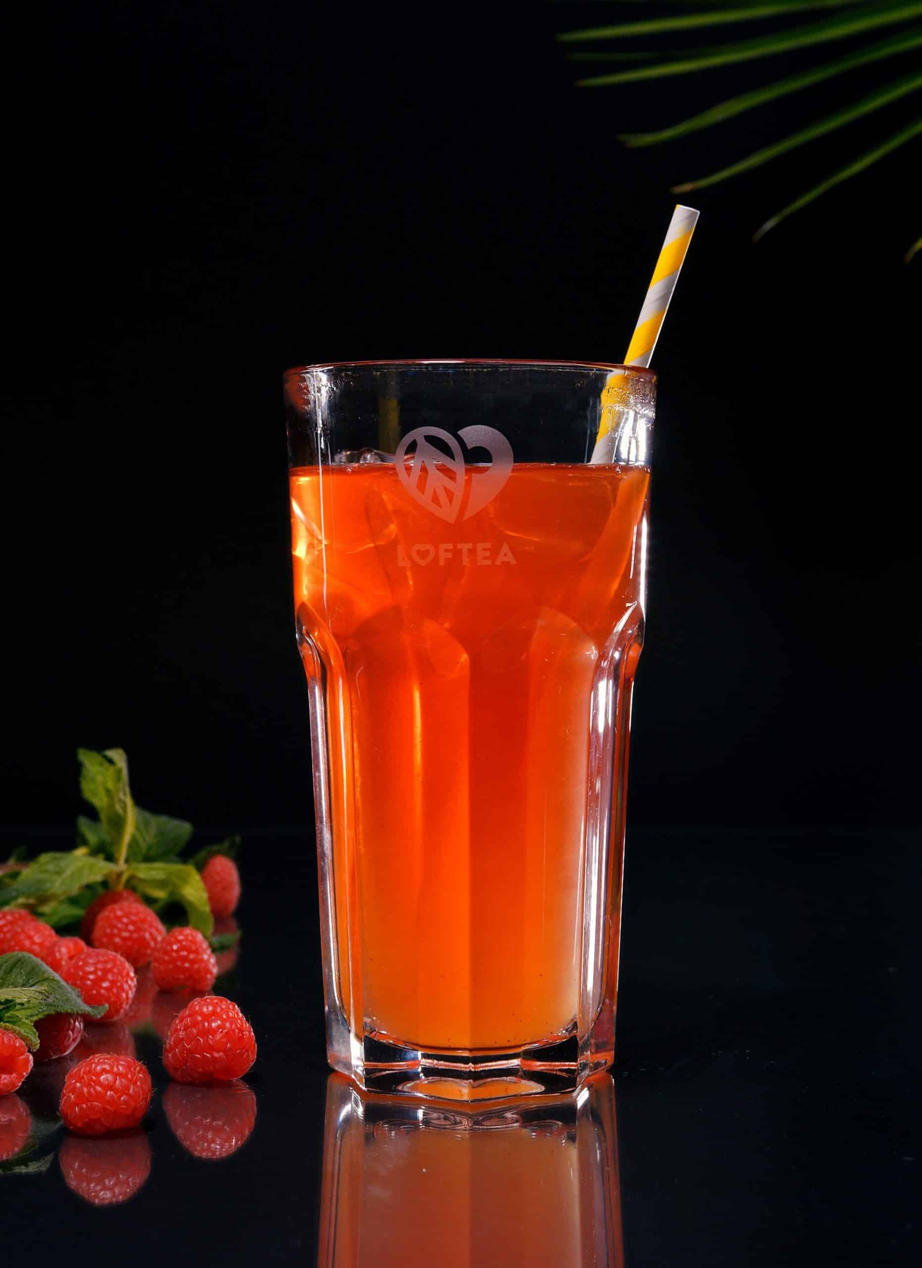 Ein hohes Glas orangefarbenen Tees mit einem gelb-weiß gestreiften Strohhalm steht vor einem dunklen Hintergrund. Kräftige Himbeeren, die an das Flair von HORST HIMBEER erinnern, und Minzblätter sorgen neben dem Glas für einen Farbtupfer. In der oberen rechten Ecke ist ein grünes Blatt zu sehen.