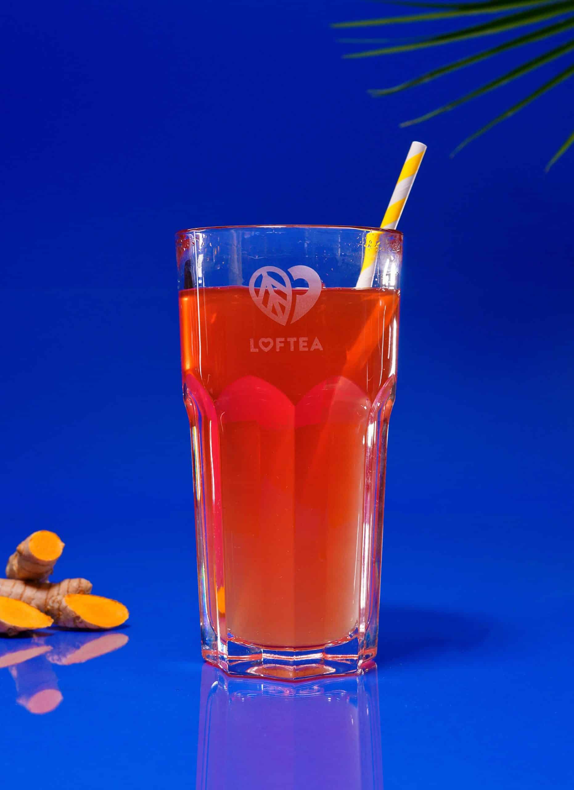 Ein großes Glas Orangentee mit einem gelb-weißen Strohhalm steht vor einem leuchtend blauen Hintergrund und verkörpert den Geist von JOGI JONTE. Daneben sind einige Kurkumawurzeln angeordnet, und aus der oberen rechten Ecke ragt elegant ein Palmblatt hervor.