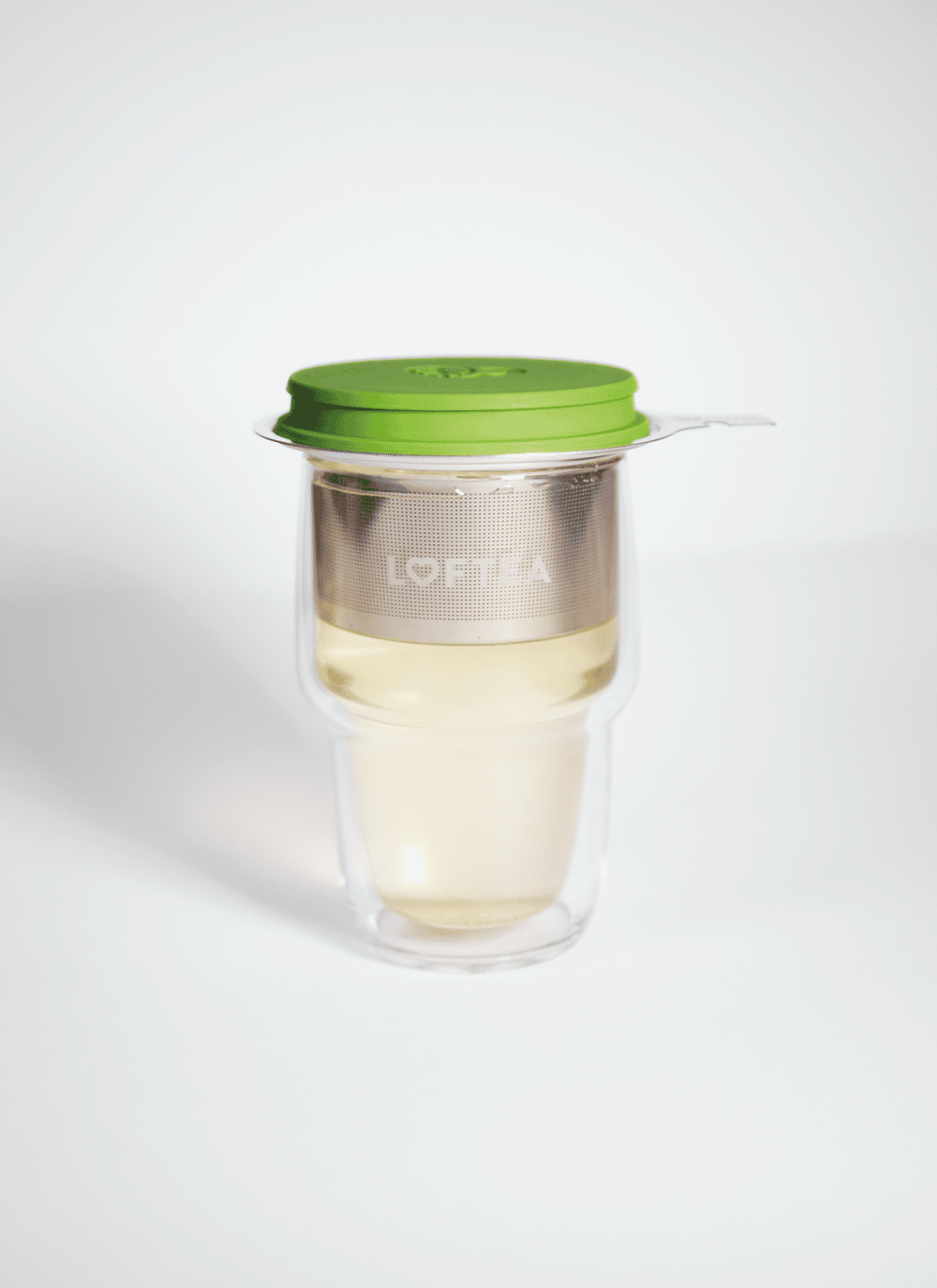 Ein transparentes LOFTEA Doppelwand-Glas enthält hellen Tee mit einem Edelstahl-Sieb und einem grünen Silikondeckel. Auf dem Sieb ist das Wort LOVEKA zu sehen, was es zu einer eleganten Ergänzung für jedes Tee-Set macht.