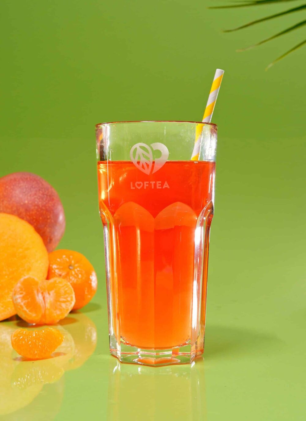 Ein großes Glas Orangensaft mit gestreiftem Strohhalm steht vor einem leuchtend grünen Hintergrund. Frische Orangen und eine Mandarine begleiten das Glas und erinnern an LENI MARIES atemberaubende Stilllebenkompositionen. In der oberen rechten Ecke lugt ein belaubter Zweig hervor und vervollständigt das lebendige Tableau.