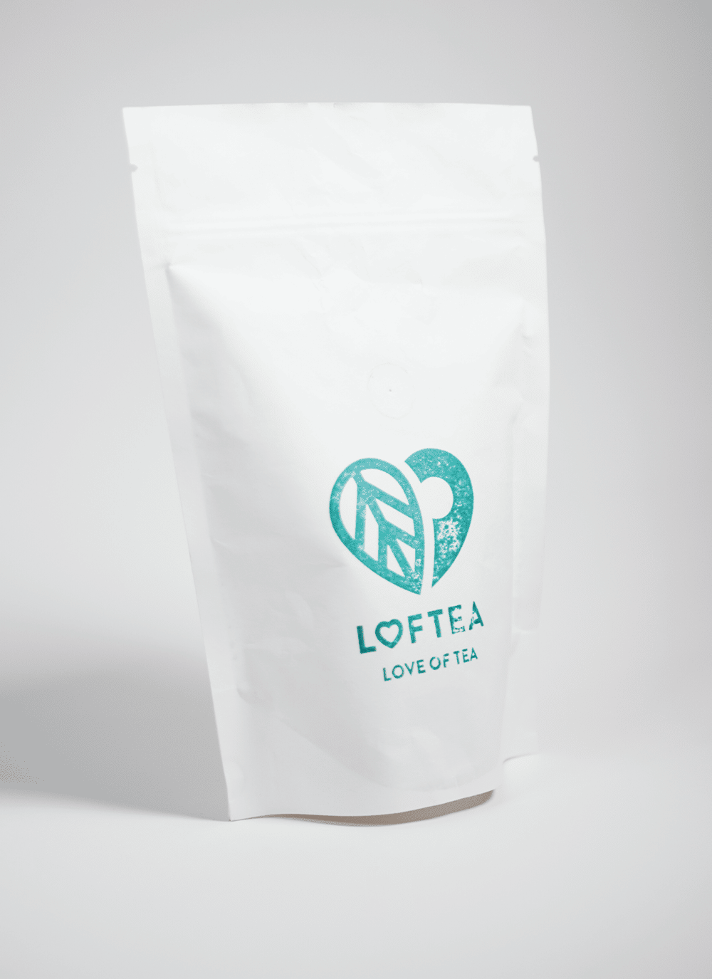 Ein weißer Teebeutel steht aufrecht vor einem hellen Hintergrund. Er zeigt ein grünes herzförmiges Logo mit Blättern und darunter die grün aufgedruckten Worte „LOFTEA LOVE OF TEA“, die darauf hinweisen, dass 200 g Tee enthalten sind.