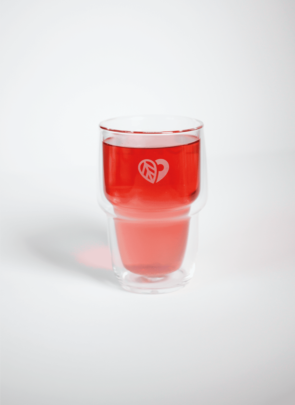 Ein Glas aus dem LOFTEA – Doppelwand-Glas Tee Set inkl. 200g Tee nach Wahl, gefüllt mit einer leuchtend roten Flüssigkeit, steht auf einer weißen Oberfläche. Es zeigt ein kleines weißes Logo mit einem Herz und abstrakten Formen.