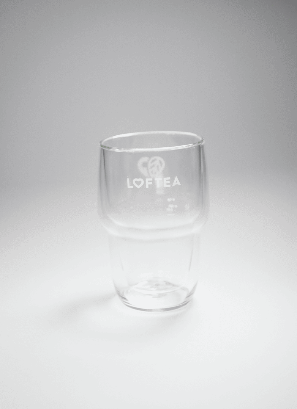 Eine Tasse aus klarem Doppelwandglas aus Borosilikatglas mit dem Wort LOFTEA und einem Herzlogo steht elegant auf einer weißen Oberfläche. Der sanft beleuchtete Hintergrund unterstreicht seine Schlichtheit und seinen Charme.