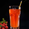 Ein hohes Glas orangefarbenen Tees mit einem gelb-weiß gestreiften Strohhalm steht vor einem dunklen Hintergrund. Kräftige Himbeeren, die an das Flair von HORST HIMBEER erinnern, und Minzblätter sorgen neben dem Glas für einen Farbtupfer. In der oberen rechten Ecke ist ein grünes Blatt zu sehen.