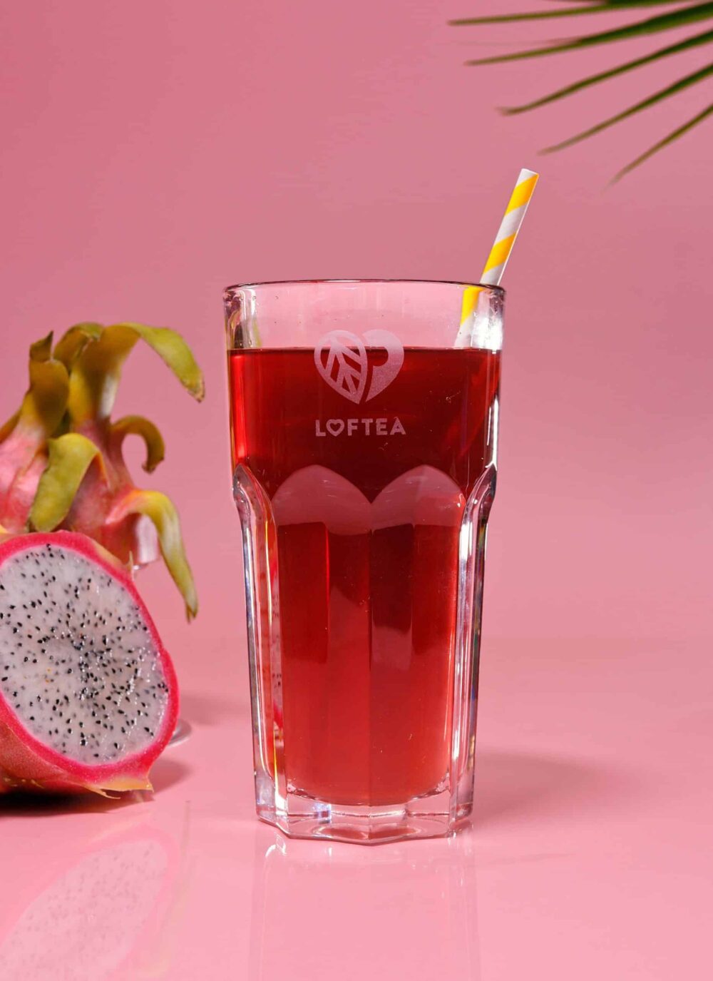 Auf einem rosa Hintergrund steht ein Glas mit rotem Eistee IDA ROSE. Auf dem Glas befindet sich ein gelb gestreifter Strohhalm und das Wort „Loftea“. Daneben sorgen eine halbe Drachenfrucht und ein Teil eines Palmblatts für eine exotische Note.