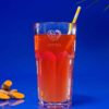 Ein großes Glas Orangentee mit einem gelb-weißen Strohhalm steht vor einem leuchtend blauen Hintergrund und verkörpert den Geist von JOGI JONTE. Daneben sind einige Kurkumawurzeln angeordnet, und aus der oberen rechten Ecke ragt elegant ein Palmblatt hervor.