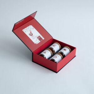 Eine festliche, offene Geschenkbox mit vier hübsch angeordneten 50-g-Gläsern Honig mit weißen Etiketten, ideal zum Weihnachtstee. Der Deckel zeigt stolz das Logo „X-MAS BOX – 4x50g Weihnachts-Tee“ und ein farbenfrohes Design, alles vor einem einfachen hellgrauen Hintergrund.