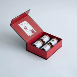 Eine festliche, offene Geschenkbox mit vier hübsch angeordneten 50-g-Gläsern Honig mit weißen Etiketten, ideal zum Weihnachtstee. Der Deckel zeigt stolz das Logo „X-MAS BOX – 4x50g Weihnachts-Tee“ und ein farbenfrohes Design, alles vor einem einfachen hellgrauen Hintergrund.
