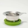 Das LOFTEA – ICE BREW Tee-Set enthält ein Teesieb aus Edelstahlgeflecht mit einer grünen Silikonbasis, die das Set perfekt ergänzt. Es verfügt über einen praktischen Griff zum einfachen Herausnehmen und präsentiert sich elegant vor einem schlichten weißen Hintergrund.