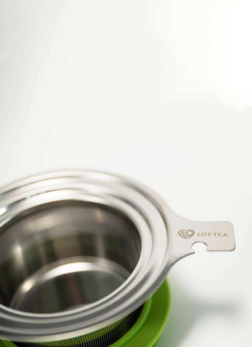 Ein LOFTEA – ICE BREW Tee-Set, komplett mit einem Teesieb aus Edelstahl mit Siebkorb und beschriftetem Griff, ruht elegant auf einer grünen Silikonbasis. Die Szene ist vor einem weichen, verschwommenen weißen Hintergrund dargestellt.