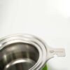 Ein LOFTEA – ICE BREW Tee-Set, komplett mit einem Teesieb aus Edelstahl mit Siebkorb und beschriftetem Griff, ruht elegant auf einer grünen Silikonbasis. Die Szene ist vor einem weichen, verschwommenen weißen Hintergrund dargestellt.