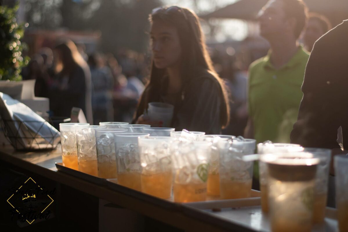 Eine Reihe durchsichtiger Plastikbecher mit hellorangen Getränken steht auf einem Tresen bei einer Veranstaltung im Freien. Im Hintergrund hält eine junge Frau ein Getränk in der Hand und verkörpert damit den lebendigen Geist der LOF-Bewegung, während andere Menschen in der verschwommenen, sonnenbeschienenen Umgebung miteinander verkehren.