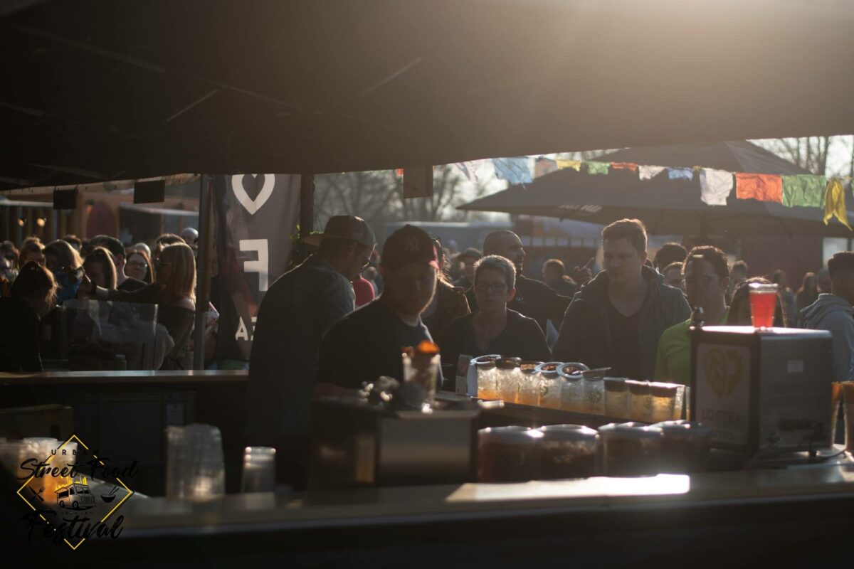 Eine geschäftige Festivalszene im Freien mit Menschen, die in die lebendige Atmosphäre der LOF-Bewegung eintauchen. Sie versammeln sich um eine Bar, auf deren Theke bunte Getränke angeboten werden. Über ihnen flattern Banner und Flaggen und fangen die Essenz eines sonnigen Nachmittags voller lebendiger Bewegungen ein.