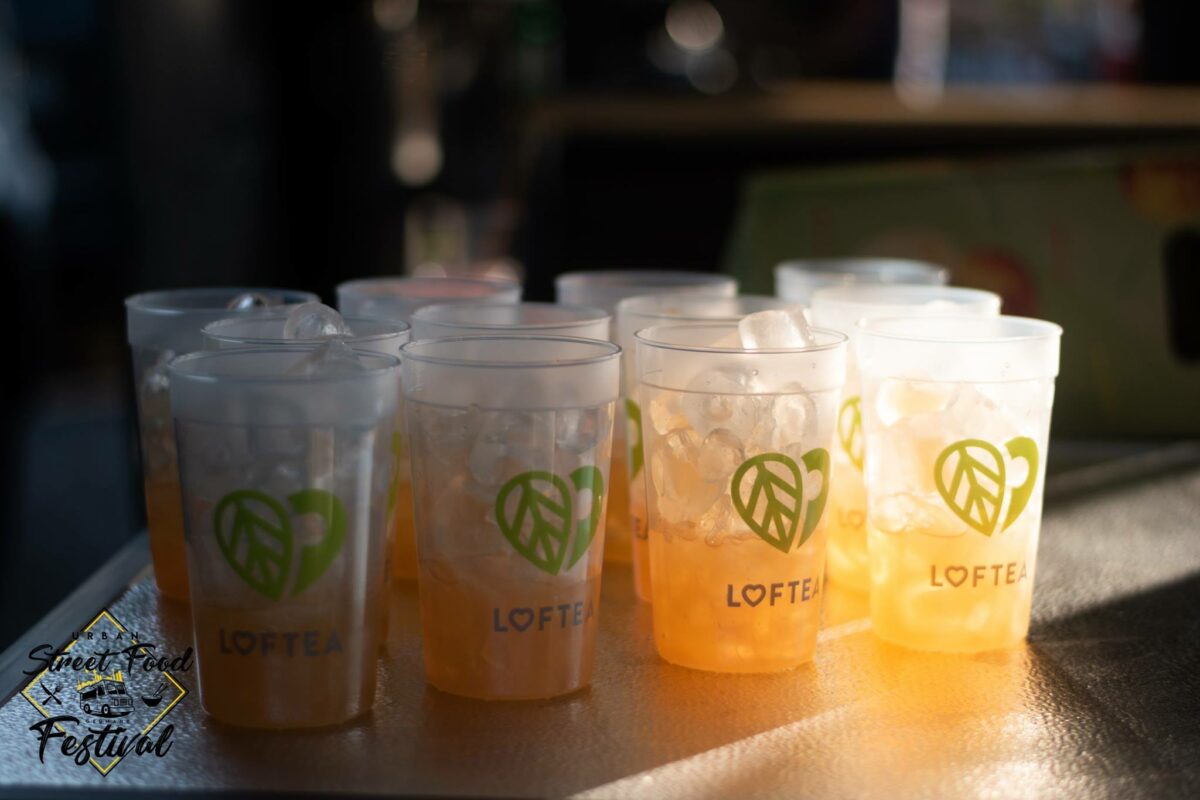 Mehrere mit Eistee gefüllte Tassen stehen auf einer Theke aufgereiht. Die durchsichtigen Tassen zeigen ein grünes Blattdesign und das Wort LOFTEA und spiegeln die lebendige Energie der Marke wider, die mit der LOF-Bewegung in Einklang steht. Das Sonnenlicht wirft einen warmen Schein und hebt das Eis in jeder Tasse hervor.