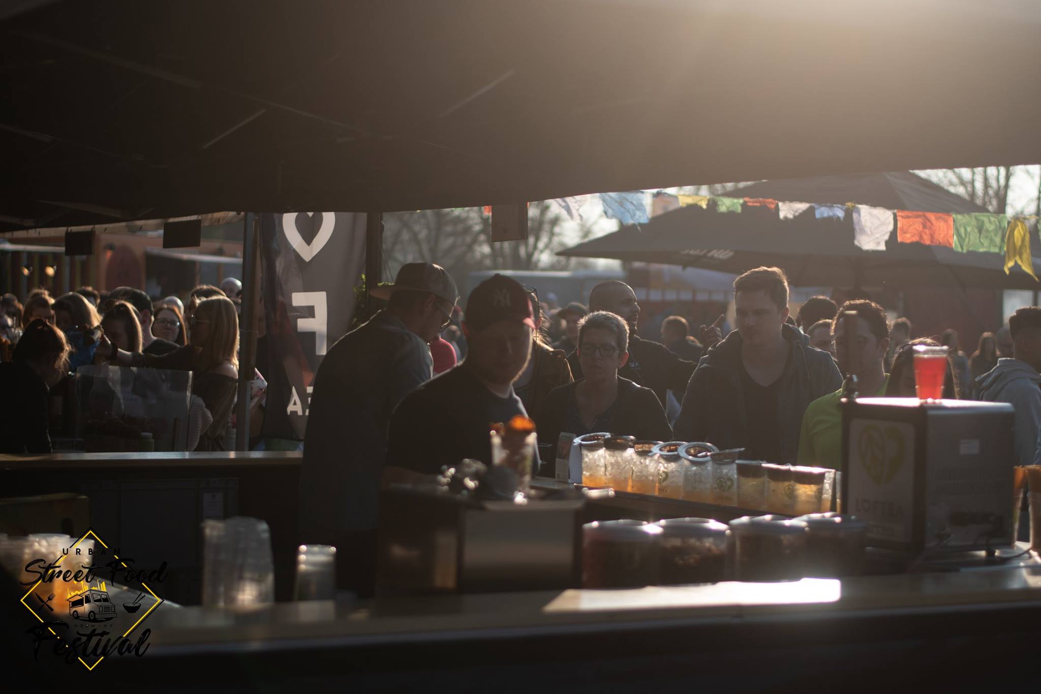 Eine geschäftige Festivalszene im Freien mit Menschen, die in die lebendige Atmosphäre der LOF-Bewegung eintauchen. Sie versammeln sich um eine Bar, auf deren Theke bunte Getränke angeboten werden. Über ihnen flattern Banner und Flaggen und fangen die Essenz eines sonnigen Nachmittags voller lebendiger Bewegungen ein.