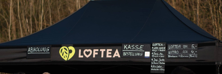 Ein schwarzes Zelt mit einem LOFTEA-Schild und einem grünen Logo sticht ins Auge. Separate Banner mit den Aufschriften „ABHOLUNG“ und „KASSE BESTELLUNG“ weisen den Kunden den Weg, während eine Getränkekarte mit Preisen zwischen 3 und 5 Euro wie Tee und Nitro auflistet und zum Erlebnis einlädt. Bäume bilden eine ruhige Kulisse für diese lebendige LOF-Bewegungsszene.